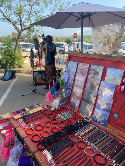 A street vendor is playing a musical instrument in San Luis De Sabinillas, Spain. The vendor, wearing a dark sleeveless shirt and brown pants, stands behind a table displaying various items for sale. The table is covered with a red cloth and features numerous CDs, dreamcatchers, necklaces, bracelets, and what appear to be wooden flutes or panpipes. The vendor is positioned under a large gray umbrella, and a microphone and speaker are set up next to them. In the background, several cars are parked, and lush green trees and foliage line the area. A sign with a red circle and a horizontal bar is visible in the distance. The sky is clear and blue, indicating a sunny day.
