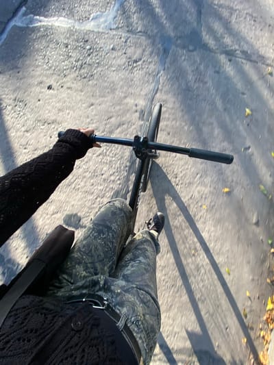 A first-person perspective shot from atop a bicycle moving along a paved surface. The cyclist's left hand grips the handlebar, clad in a black knit sleeve. The bike's front wheel and handlebars are visible, casting shadows on the pavement. The cyclist's legs, covered in camouflage pants, are visible to the right. A black shoe is visible on the right foot. A black bag is slung across the torso. The surface appears to be concrete, possibly a sidewalk or bike path. The location is presumed to be Monte Grande, Argentina, but there are no local landmarks or identifiers visible in the frame to confirm this. No other people, subjects, objects, or actions are visible in the frame. The environment appears to be outdoors during daylight hours, with dappled sunlight and shadows.