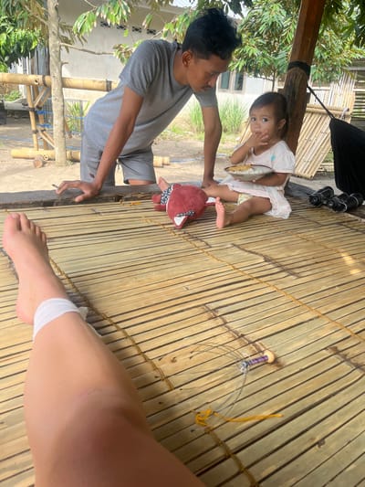 The scene is set outdoors on a raised platform with a light-colored, woven bamboo mat floor in Desa Batu Layar, Indonesia. In the immediate foreground, the bare legs of an individual are extended, with a white fabric bandage wrapped around the right ankle. To the right, an adult male, dressed in a grey t-shirt and grey shorts, leans forward onto a wooden beam that forms the edge of the platform. He holds a lit cigarette in his right hand and looks down towards a young child. The child, seated cross-legged on the bamboo mat, wears a light-colored dress and is eating from a small, light-colored bowl containing rice and another food item, with her right hand near her mouth. Adjacent to the child, a red fox-shaped plush toy lies on the mat. Further to the right, a pair of black binoculars rests near a dark, folded hammock. In the lower-middle foreground, a fishing rig composed of a purple-wrapped spool of line, hooks, and a yellow line is visible on the bamboo mat. The background features green foliage from trees, a wooden structure made of bamboo poles, and a distant light-colored building with a dark roof. The ground beneath the platform appears to be dirt.