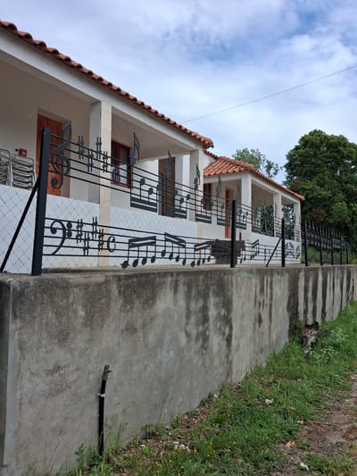 A low-angle, color photograph depicts the exterior of a two-story building in Soure, Portugal. The building features a white façade with a red-tiled roof. The railing is decorated with black ironwork musical notes and staff lines. The building has multiple evenly spaced windows with visible red frames. The building sits atop a gray concrete wall. Beyond the wall, a strip of grass separates the wall from the ground. The sky is overcast with a few patches of blue visible. A tree is visible above the building. Power lines are seen near the left edge.