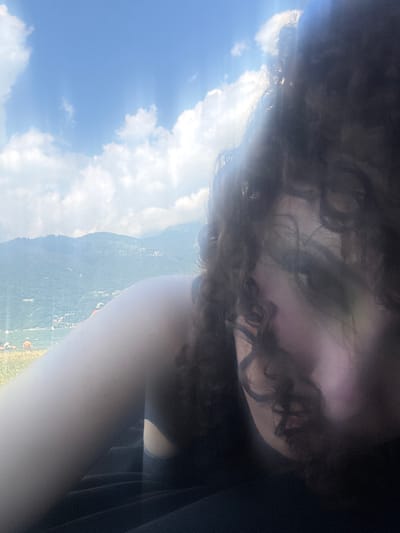In a close-up, low-angle selfie, a person with dark, curly hair is positioned in the foreground, their face partially obscured by their hair and the bright sunlight. Their left arm is visible, extending diagonally across the frame from the lower left. In the background, a picturesque view of a body of water, likely Lake Como, is visible, with lush green mountains rising behind it under a bright, partly cloudy sky. The scene appears to be set in or near Colico, Italy, given the prominent mountainous landscape and proximity to the lake. The overall atmosphere is one of casual enjoyment of the outdoors.