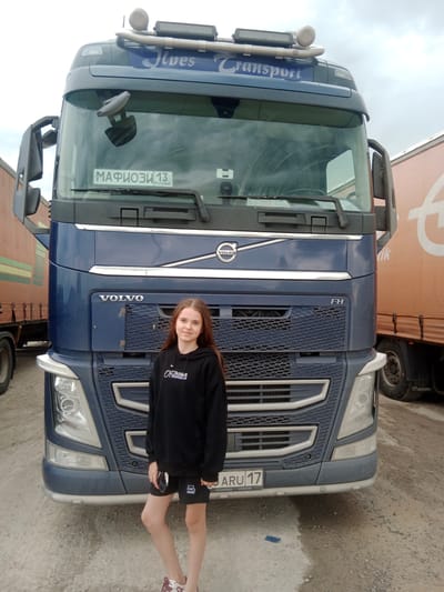 A young woman with long, brown hair stands in front of a blue Volvo FH semi-truck. She is wearing a black hoodie with white lettering and black shorts. The truck has "JIVES TRANSPORT" written on the top of the windshield and the "VOLVO" logo on the front grill. A license plate is visible on the windshield with the inscription "МАФИОЗИ 13" and "RUS". Another license plate on the lower front bumper reads "ARU 17 VOLVO".
Behind the Volvo, the side of an orange semi-trailer truck with a large white logo is visible. The trailer has a "KRONE" marking at the bottom. The ground is a mix of gravel and dirt, and the sky is overcast, suggesting a cloudy day. The overall scene appears to be a truck stop or industrial area. The woman is looking directly at the camera with a slight smile. The time of day is not explicitly indicated, but the lighting suggests it is daytime. The location is identified as Sarytogay, Kazakhstan.