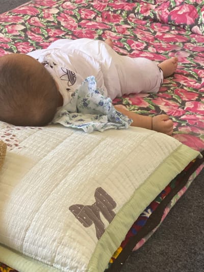 The image shows a close-up, high-angle view of a baby sleeping on a floral patterned sheet. The baby is lying on its stomach, with its head turned to the left. The baby is wearing a white outfit with a blue floral pattern on the collar. In the foreground, there is a light green quilted blanket with a brown bow applique. The background is a dark grey surface, possibly a carpet or floor. The scene appears to be indoors, and there are no specific landmarks or features to indicate its location in Arbagish, Uzbekistan.