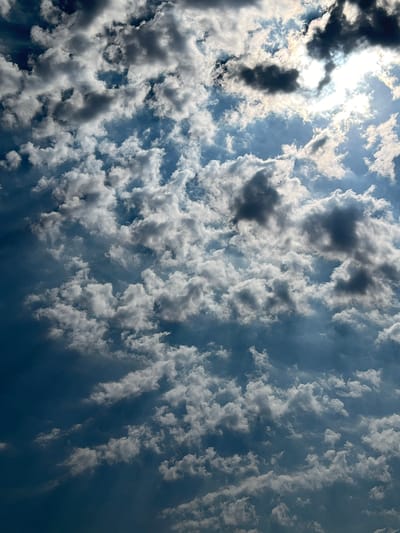The image is a close-up, vertical shot of a dynamic sky filled with a variety of clouds. The clouds are predominantly white and gray, with some darker, more ominous formations interspersed. The sun is visible in the upper right quadrant, partially obscured by clouds, casting a bright, diffused light and creating rays that penetrate the cloud cover. The sky transitions from a bright blue near the sun to a deeper, darker blue towards the bottom of the frame, suggesting depth and atmospheric perspective. The overall impression is one of a dramatic and possibly changing weather condition. There are no people, subjects, objects, actions, interactions, or any identifiable landmarks of Ringsted, Denmark, visible in the image. The scene is entirely focused on the sky.