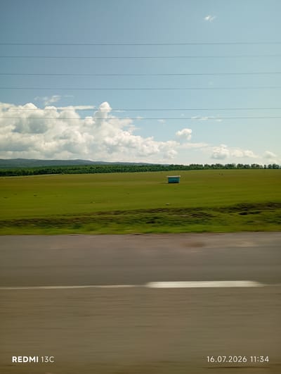 A wide, scenic view captures a lush green field under a bright, partly cloudy sky in Galbai, Russia. A long, dark road stretches across the foreground, blurred due to motion. In the middle ground, a solitary, small, light blue structure with a dark roof stands in the expansive grassland. A line of dense, dark green trees forms a distant horizon, with rolling hills visible beyond them. Several thin, dark power lines traverse the sky, and a few scattered dark specks, possibly livestock, are visible in the field. The image is watermarked with "REDMI 13C" and the date "16.07.2026 11:34" in the bottom corners.