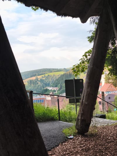 The image captures a view through the opening of a rustic, wooden structure, likely a lookout or pavilion, in Triberg im Schwarzwald, Germany. The foreground is dominated by the dark, rough texture of large wooden beams that frame the scene. To the left, a dark, textured beam of wood is prominent, and to the right, another thick, weathered tree trunk leans into the frame.
Beyond the wooden structure, a picturesque landscape unfolds. Rolling green hills dotted with evergreen forests form the middle ground, transitioning into a town nestled in the valley. The town features red-tiled roofs, buildings with traditional architecture, and a prominent white church steeple. A black rectangular sign is mounted on a pole in the middle ground, partially obscured by the tree trunk. Solar panels are visible on the roof of a building in the town. A metal railing runs along the edge of a pathway in the middle ground, which appears to be paved with gravel and is bordered by lush green grass and some wildflowers. The sky above is a mix of white clouds and blue, suggesting an overcast but bright day. The ground in the immediate foreground is covered with wood chips.