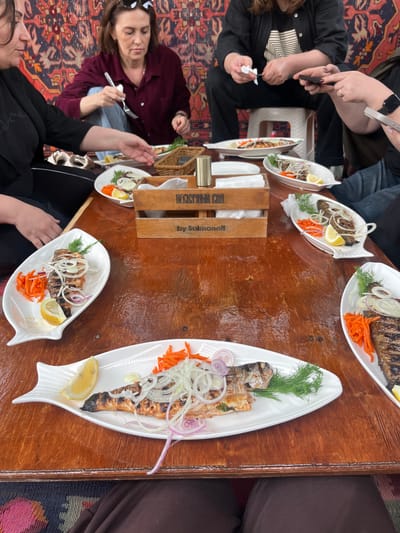 A group of people are seated around a wooden table laden with plates of grilled fish in what appears to be a restaurant or dining setting. The fish dishes are served on white, fish-shaped plates and garnished with lemon slices, shredded carrots, dill, and onion rings. A wooden box with the words "HECKYPHHый COA" and "by Salmanoff" in Cyrillic text is placed centrally on the table. People are interacting, with some holding forks and others with napkins or phones in their hands. The setting is inside, with an ornate carpet visible under the table, suggesting a location that may be in Miatli, Russia, based on the Cyrillic text.