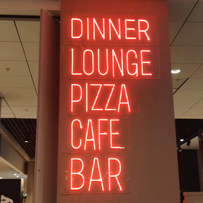 The image displays a vertical arrangement of five red neon signs mounted on a light brown column within an interior commercial environment in Stockholm-Arlanda, Sweden. From top to bottom, the illuminated signs spell "DINNER," "LOUNGE," "PIZZA," "CAFE," and "BAR." Each word is formed from red neon tubing and affixed to a transparent rectangular acrylic panel, which is secured to the column by four visible corner fasteners. The background on both sides of the column features a dropped ceiling with light-colored acoustic tiles, dark grid lines, and integrated recessed lighting fixtures. The left background additionally reveals a lower section with a darker, slatted ceiling, more lighting elements, and a partial white arrow sign. The right background shows similar ceiling details and distant dark architectural elements. No individuals are present in the scene.