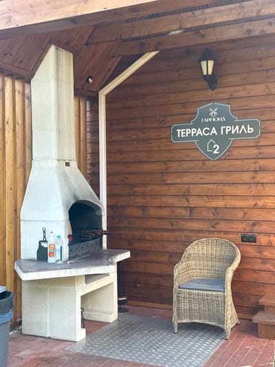 This image depicts an outdoor covered patio area, likely part of a restaurant or entertainment venue, situated in Verkhnee Myachkovo, Russia. The focal point on the left is a large, white, concrete barbecue grill with a cooking grate and skewers of meat inside. The countertop of the grill area holds several cleaning supplies and a small box of matches. To the right of the grill is a woven wicker armchair with a grey cushion. Behind the armchair, mounted on a dark wooden plank wall, is a sign with a dark green shield shape. Inside the shield, in white lettering, is the word "ГАРМОНД" at the top, followed by "ТЕРРАСА ГРИЛЬ" below it, and the number "2" in a smaller shield at the bottom. A black wall-mounted lantern with a lit bulb is positioned above the sign. The ceiling is constructed of dark wooden planks, and the walls are also made of similar wooden planks. An electrical outlet with two sockets is visible on the wall near the chair. The ground is a mix of brick paving and a metal diamond plate mat. A dark trash can is partially visible on the far left. The overall impression is a clean and functional outdoor space designed for grilling and relaxation.