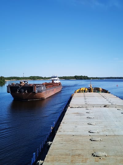 The image depicts a clear, sunny day on a wide inland waterway near Kargulino, Russia, with two barges as primary subjects. The perspective is from the deck of a flat-topped barge, which occupies the lower and right portions of the frame. Its deck is a textured grey, featuring numerous evenly spaced circular hatches. A distinctive blue railing runs along the visible port side of this foreground barge.
Another, rust-colored barge with a dark grey upper deck and a white superstructure at its stern is actively sailing past, moving from left to right across the frame. The number "59221" is clearly visible on the white cabin of this passing vessel. Its movement generates noticeable ripples and a wake in the deep blue water.
The waterway extends into the distance, flanked by lush green forests on both banks. A solitary red buoy is visible further down the waterway. The sky above is a brilliant, cloudless blue, indicating bright, clear weather, likely midday given the strong illumination. The scene conveys a sense of peaceful transit on a busy shipping route. No individuals are visibly present in the image.
