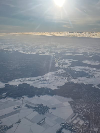 An aerial photograph captures a snow-covered winter landscape, likely taken from an aircraft. The sun, prominent in the upper right, casts intense flares and streaking rays across the scene. The sky above is clear blue, while a distinct, elongated band of stratified clouds spans the distant horizon, approximately one-third of the way up from the bottom edge. A faint contrail is visible in the upper left quadrant.
The terrain below is extensively covered in snow, appearing white and textured, with darker areas indicating exposed land or dense forestation. A prominent river, the Isar River, flows from the top-middle of the frame towards the bottom-right, visible as a darker, winding line against the snow. Parallel to the Isar River on its western bank, a major highway (likely Bundesstraße 11 or Autobahn 92) traverses the landscape, appearing as a bright, linear feature reflecting sunlight.
The city of Freising, Germany, is identifiable in the bottom-right quadrant of the image as a sprawling urban area with a high density of buildings and a discernible street network. Buildings and rooftops within Freising are covered in snow. Smaller villages and hamlets, characterized by clusters of structures amidst snow-covered agricultural fields, are scattered throughout the mid-ground and foreground. Patches of dark green/brown forests punctuate the landscape, contrasting with the white snow. No individuals are discernible within the frame due to the high altitude.