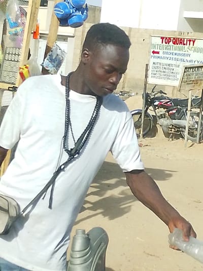 A young man with dark skin and a short, faded haircut is captured outdoors in bright daylight. He is wearing a plain white short-sleeved t-shirt and multiple dark-colored necklaces or prayer beads around his neck, some of which extend down his chest with dark, possibly leather, tassels or straps. His gaze is directed downwards and to his right, focusing intently on something below him or in his hands.
His right arm is extended downwards, and he is holding a clear, textured plastic bottle. Near his left side, a grey, possibly plastic, container or flask with an open top is visible. To the left of the man, various items, including blue children's shoes and plastic bags containing small goods, are hanging from what appears to be a makeshift stall or stand.
In the background, to the right of the man, a prominent white sign with red text is visible. The text reads: "TOP QUALITY INTERNATIONAL SCHOOL," followed by "CRECHE NURSERY," and "PRIMARY J.S.S ISLANIYYA." Below this, the motto "LEARNERS TODAY, LEADERS TOMORROW" is displayed. Further details on the sign include "MR. NERMANU/GULUMALGA," and "KANO TEL: 08036361251, 09035230937/08020428347." Partially visible next to this sign is another smaller sign that reads "DAYYANIY LAUNDRY SERVIC."
Behind the signs, several motorcycles are parked on a light brown, dusty, or sandy ground. Light-colored buildings are visible in the distance under a bright sky. Distinct shadows are cast on the ground by the man and other objects, indicating bright sunlight, likely during the midday or early afternoon. The scene suggests an outdoor commercial or street market setting, possibly in Kano, Nigeria, given the phone numbers on the school sign. The man appears to be engaged in an activity related to the items he is handling, possibly a transaction or preparation for sale.