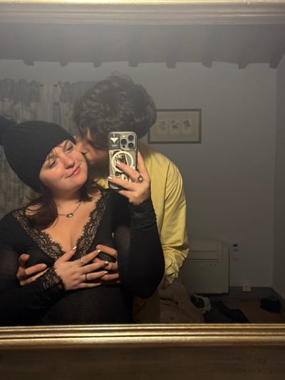 A mirror selfie captures a couple. The person on the left, with curly dark hair, wears a yellow shirt and embraces the other. The person on the right, wearing a black knit hat and a black lace-trimmed top, is holding up the phone. Visible in the background is a framed picture on the wall, a white air conditioner, and a bed with rumpled bedding. The overall tone of the image is intimate and casual.