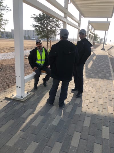 The image displays an outdoor scene in Hanak, Saudi Arabia, featuring three adult males on a paved walkway. One male is seated on a white metal bench structure, wearing a grey baseball cap, a grey shirt, and a yellow high-visibility vest; he has a mustache and beard and is looking forward. Another male stands with his back mostly to the camera, dressed in a grey baseball cap, a black hooded jacket, and dark trousers. A third male stands to the right of the second, facing partially left, also wearing a grey baseball cap, a black hooded jacket, and dark trousers; he also has a mustache.
The ground consists of interlocking grey pavers, casting strong shadows from the individuals and the overhead white metal shade structures. To the right of the paved area, an asphalt road is visible. To the left, an unpaved, arid plot with sparse vegetation and gravel extends into the background, where a large, multi-story building with numerous windows is present under a clear sky.