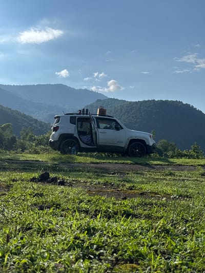 A white Jeep Renegade is parked on a dirt road with its driver's side door open. The vehicle is on a grassy overlook with a panoramic view of lush green mountains and a blue sky with scattered clouds. The foreground shows dew-covered grass and some dark soil clumps. The Jeep has items on its roof rack and a water bottle is visible inside the vehicle. The scene suggests an outdoor adventure or camping trip in a mountainous, rural location. It is not possible to determine the location within the city of Chkvaleri, Georgia from the image alone.