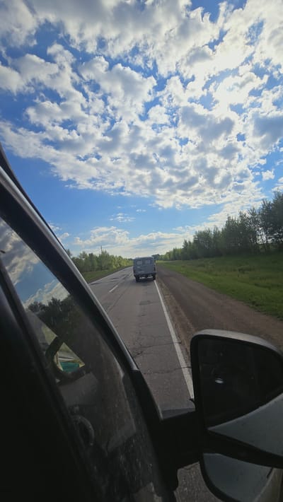 The image depicts a view from inside a vehicle traveling on a paved road. The road is cracked and has a white dividing line. A grey van is visible driving ahead on the same road. The right side of the road is bordered by a grassy area and a line of trees. The sky is bright blue with scattered white clouds, and the sun is visible through some of the clouds. The left side of the image shows a portion of the interior of the vehicle, including the side mirror and window, which reflects the sky and trees. The scene appears to be in a rural or suburban area. No specific location within Кама-Исмагилово, Russia is identifiable from the image alone.