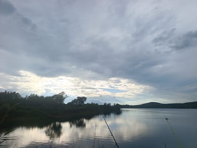 The image depicts a wide, calm body of water, likely a lake or reservoir, with a tree-lined shore on the left and rolling hills in the distance. The sky is filled with dramatic, layered clouds, with patches of sunlight breaking through on the left side, illuminating the water and the trees. Two fishing rods are visible in the foreground, angled into the water, suggesting a fishing activity. The overall atmosphere is serene and somewhat overcast, hinting at either dawn or dusk. The location is identified as Yaroslavsky, Russia, implying a natural landscape setting near this city.
