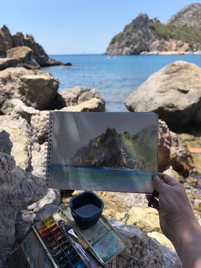 A hand holds a spiral-bound sketchbook with a watercolor painting of a rocky coastline and blue water. The painting depicts a sunlit, rugged cliff face with patches of green vegetation and the sea below. To the left of the sketchbook is a watercolor paint palette with various colors, two paintbrushes, and a black container filled with water, all resting on a rocky surface. The background is a blurred view of a rocky shoreline and the Mediterranean Sea under a clear blue sky, with distant mountains and a few boats visible. The scene is set in the city of Datça, Turkey, a coastal town known for its natural beauty and historical sites.