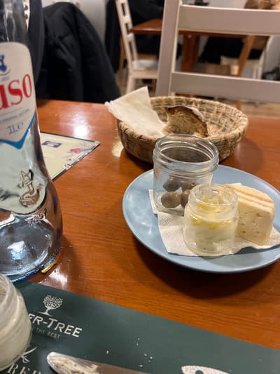 A light blue plate holds a piece of cheese, a small jar of pickled onions, and a jar of olives, all resting on a white napkin. Next to the plate, a woven basket contains bread and a white cloth. The scene is set on a wooden table, partially covered by a green menu or placemat. A glass bottle with a blue and white label is visible in the foreground, along with a small glass jar. A white chair frame and a glimpse of clothing are in the background.