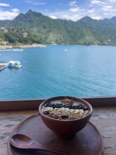 A breakfast bowl sits on a wooden surface with a mountain and ocean view. The bowl appears to contain yogurt, granola, banana slices, and chocolate chips. It sits on a terracotta plate next to a wooden spoon. In the background, a mountain descends into a body of water. The day is bright with a few clouds in the sky.