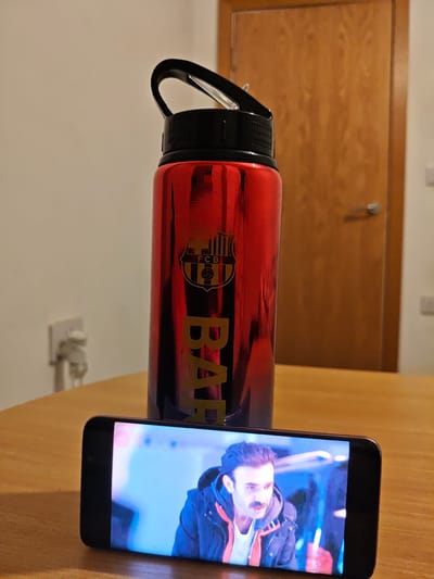 The image displays an indoor scene in Swindon, United Kingdom, with a focus on items arranged on a light brown wooden table. In the foreground, a black smartphone is positioned horizontally, actively displaying video content. The screen shows a man with dark hair and a prominent mustache, wearing a dark hooded jacket with an orange or red interior lining over a white shirt, set against a blurred blue background. Immediately behind the smartphone, a tall, reflective red metallic water bottle stands upright. This bottle features a black screw-top lid with an integrated black carrying loop and is emblazoned with a gold-colored FC Barcelona crest and the vertical word "BARÇA". The background features a plain light-colored wall. To the right, a light brown wooden door with a silver handle is visible. Two white wall-mounted electrical sockets or switches are present: one on the left side of the wall and another on the right, near the door frame.