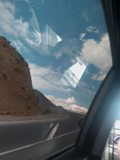 The image captures a dynamic view from inside a vehicle, looking out a side window at a landscape in Tigranashen, Armenia. The original photo is significantly rotated counter-clockwise.

When oriented upright, the scene reveals a bright blue sky filled with scattered white, fluffy cumulus clouds. Below the sky, a brown, arid, and rocky hillside or mountain slope occupies the mid-ground. An asphalt road with clear white markings – including a double solid line and a single dashed line – curves alongside the terrain. A metal guardrail can be seen bordering the road in sections.

The most striking aspect of the image is the strong reflection on the car window, which overlays the external view. Reflections of the sky and clouds are prominently visible, creating a layered effect. Parts of the vehicle's interior are also reflected, including a section of a dashboard, possibly a vent, and some illuminated controls in the upper right portion of the image. Ambiguous shapes in the reflections might suggest the presence of occupants or interior structures, but no distinct people are visible.

The scene is bathed in bright daylight, indicating clear and sunny weather with some clouds. The rugged landscape and road suggest travel through a mountainous or hilly region. No discernible text is visible within the image.