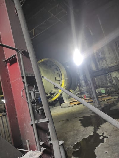 The image depicts the interior of an industrial facility, possibly a cement plant or similar manufacturing site, located in the city of Kyzyl, Kazakhstan. Dominating the scene is a large, cylindrical industrial machine, possibly a rotary kiln, with a yellow band around its circumference. In the foreground, a thick, red metal beam supports a grey pipe. The floor is concrete and wet in patches, indicating a humid or recently cleaned environment. Light sources, including a bright overhead light, cast strong reflections and lens flares across the image, obscuring some details. The overall atmosphere is dimly lit and industrial, with exposed pipes, electrical boxes, and various metal structures visible. There are no people present in the frame.