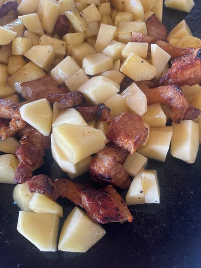 The scene, located in Petrivka, Ukraine, presents a high-angle, close-up view of cooked food. The image primarily features numerous irregularly diced pieces of potato and seared meat. The potatoes are predominantly light yellow to off-white, cut into various polygonal shapes, exhibiting some minor surface browning and a slight oily sheen. Interspersed among the potatoes are chunks of dark reddish-brown meat, characterized by visibly seared surfaces, darker charred edges, and a glistening appearance indicative of rendered fat. These meat pieces vary in size and shape, from elongated strips to more compact cubes. The food rests on a dark, non-reflective surface, likely a cooking pan or skillet, with some isolated dark spots visible on the surface. No individuals or further environmental details are discernible.