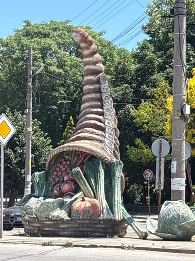 This image depicts a large, colorful statue of a cornucopia overflowing with fruits and vegetables. The statue is situated outdoors, likely in a public space within the city of Isperihovo, Bulgaria, given the context. Dominating the foreground is the intricately sculpted cornucopia, made of what appears to be concrete or a similar material, painted to resemble a woven basket. Inside the horn, various produce items are visible, including red berries (possibly grapes or cherries), tomatoes, carrots, and what look like leeks or green onions. The base of the statue is adorned with more vegetables, such as cabbage and what appears to be a pumpkin.

In the background, lush green trees form a dense canopy, suggesting a well-treed area or park. A clear blue sky is visible above the trees. To the right of the statue, a utility pole with various wires and signage is present. One sign, partially visible, appears to be a traffic sign. Further to the right, another utility pole and more signage are visible, along with some yellow foliage from a tree. To the left of the statue, a yellow diamond-shaped road sign with a black border is mounted on a pole. A portion of a car, with a license plate reading "PAS", is visible in the lower left corner of the image. The overall scene suggests a warm, sunny day in an urban or suburban environment, showcasing a public art installation celebrating agriculture or abundance.