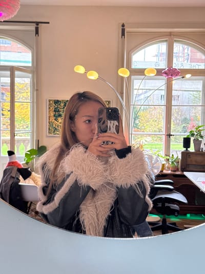 A person is observed taking a mirror selfie indoors, in a residential or office setting located in Wallisellen, Switzerland. The individual, possessing light brown hair and subtle white adornments on the right side of their face, holds a silver iPhone featuring a black and white patterned case, obscuring the majority of their face. They are dressed in a black jacket with prominent light beige/pinkish faux fur trim on the collar and cuffs, worn over a black long-sleeved garment.
The reflective surface reveals a room with light-colored walls and two large arched-top windows. Through the windows, an autumnal exterior is visible, characterized by trees displaying yellow and orange foliage, a stone balustrade, and sections of distant buildings.
The interior features a modern floor lamp with five flexible arms, four terminating in yellow, spherical lights, and one in a distinct pink, cloud-shaped light. Additional objects within the reflected space include a black office chair, a brown wooden cabinet supporting an antique-style radio and a black speaker, several potted plants, a white oscillating fan, and a partially visible white feathery pendant light fixture. A green ambient light source is present near the floor, and a white chair with draped clothing is visible on the left side of the reflection.