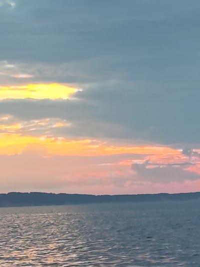 The image depicts a serene sunset over a body of water, likely a lake or sea, with a silhouetted shoreline in the distance. The sky is a gradient of colors, transitioning from a bright yellow and orange glow near the horizon, indicating the setting sun, to soft pinks and blues further up. Wispy clouds are scattered across the sky, catching the light and adding texture to the scene. The water is calm, with gentle ripples reflecting the colors of the sky. The shoreline appears as a dark, undulating line, suggesting hills or trees. The overall atmosphere is peaceful and tranquil. The location is identified as Galovo, Russia. There are no people or specific objects visible, making it a natural landscape.