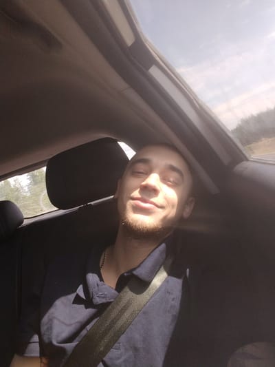 The image depicts a young man sitting in the back seat of a car, with his eyes closed and a slight smile on his face. Sunlight streams in from an open sunroof, illuminating his face and casting shadows across his body. He is wearing a dark blue polo shirt and a seatbelt is visible across his chest. The interior of the car is dark, with the headrest of the seat behind him visible. Outside the car, trees and a bright sky can be seen through the windows, suggesting a rural or suburban environment. The location is not specified as Perovo, Russia; this detail is not present in the image.
