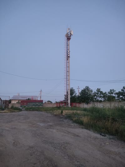 The image captures a scene in Dangir, Uzbekistan, featuring a cell tower dominating the frame against a pale blue sky. The tower, a tall, slender structure with a ladder running up its side, is equipped with multiple antennae and communication equipment at its apex. In the foreground, a dirt road leads towards a low, concrete wall topped with a green and red striped fence. Behind the fence, several buildings are visible, including a house with a red metal roof and a smaller structure with a rusty metal facade. Lush green vegetation, including trees and tall grass, grows along the roadside and behind the fence. Power lines crisscross the sky, adding to the urban infrastructure elements present in the landscape. The overall impression is of a somewhat rural or suburban area on the outskirts of the city, with a blend of natural and man-made elements.