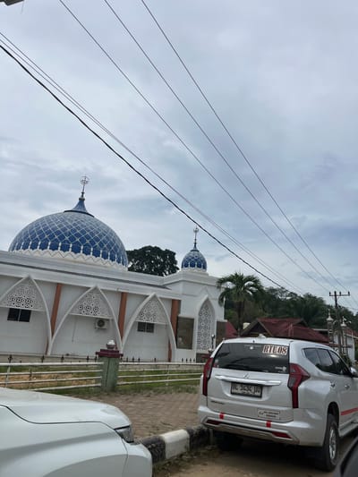 In Rantau Pauh, Indonesia, a white SUV is parked on a paved area in front of a white mosque. The mosque features a large blue dome adorned with white geometric patterns and a smaller dome on an adjacent structure. The building has white walls with decorative arched windows and orange trim. Overhead, multiple dark power lines stretch across the overcast sky. The SUV, identified by its license plate "BL 1434 UU", has a sticker on its rear window that reads "RTU 08". A white car is partially visible in the foreground, to the left of the SUV. The environment includes a metal fence bordering the mosque's grounds, and various trees and foliage in the background.