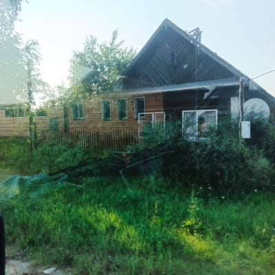 The image shows a rural scene, likely a residential property, under a bright, possibly hazy sky. The primary focus is a wooden house, constructed with horizontal wooden planks for the main body and vertical planks for the gabled roof portion. The house features several windows, some with white frames. To the right of the house, there is a satellite dish and an electrical box mounted on the exterior wall. A wooden fence with vertical pickets runs in front of the house, partially obscured by lush green vegetation and tall grass in the foreground. Trees and foliage are visible behind and to the sides of the house. The scene appears to be taken from a moving vehicle, as indicated by a slight blur and reflection on the left side of the image. The environment is consistent with a rural setting in Kambarka, Russia, characterized by traditional wooden architecture and abundant greenery. There are no people or discernible actions taking place in the image.