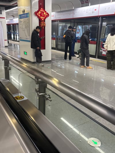 People wait at a subway station platform in front of a train. A man stands by a column looking at his phone; a red ornamental symbol adorns the column. Three individuals stand near the open train doors. A pedestrian signal with a green arrow is visible on the ground. A metal handrail runs along the platform, partially obscuring the view. The platform has a reflective surface.