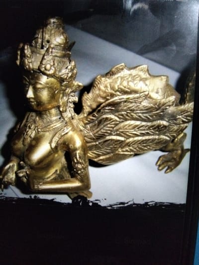 The image shows a close-up, side-view of a golden statue. The statue depicts a mythological creature with the upper body of a human female and the lower body of a dragon or similar reptilian creature. The female torso is adorned with jewelry and the head wears an elaborate crown. The creature has feathered wings and clawed feet. The statue is positioned on a white surface, with a dark, out-of-focus background. The image is grainy and has poor lighting. The statue's location in Kualaraja, Indonesia is not discernible from the visual information provided.