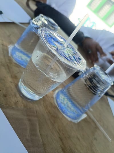 In Dayahbaro, Indonesia, three plastic cups filled with clear liquid and clear plastic lids are arranged on a wooden surface. A white straw is visible in one of the cups. The cups have blue printed designs on them. In the background, parts of two individuals are visible; one person is wearing a white shirt and has dark hair, while the other is wearing a dark garment. The scene appears to be outdoors with natural light.