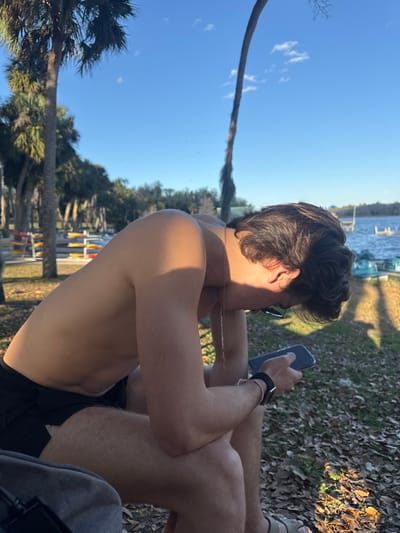 The image displays an outdoor scene in a waterfront area of Micanopy, United States. A young adult male is depicted seated, facing left with his upper body angled downwards, focused intently on a smartphone held in both hands. He is shirtless, wearing dark-colored shorts, a thin gold-colored chain necklace, and a dark watch on his left wrist. Direct sunlight illuminates his right shoulder and arm, casting distinct shadows. The ground directly beneath him consists of sparse grass and scattered dry leaves. In the background, a row of multiple kayaks or canoes, colored red, yellow, and blue, are visibly stacked on storage racks near the edge of a calm body of water, likely a lake or pond. Several tall palm trees and other deciduous trees frame the scene under a clear, bright blue sky with minimal cloud cover. A wooden pier or dock structure is partially visible extending over the water on the right side of the frame.
