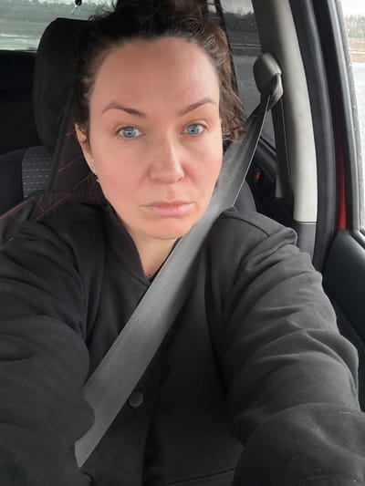 A person is sitting in a car, wearing a seatbelt and a black hooded jacket. The interior of the car is visible, including the dashboard and seats. The background shows a blurry view of the outside, possibly indicating movement or that the car is parked and it is raining. Location: Sidorovy Gory, Russia is not knowable based on image.