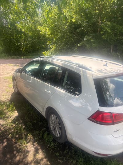 The image shows a white Volkswagen Golf station wagon parked on a dirt path surrounded by lush green trees and grass. The car is angled, with its rear and side visible. The sun is shining brightly, casting dappled shadows from the trees onto the car and the ground. The car appears to be dirty, suggesting it has been driven on unpaved roads.
The scene is set in a natural, outdoor environment, likely a rural area given the presence of trees and the unpaved path. The lighting indicates it's daytime, possibly late morning or early afternoon due to the strong sunlight and shadows. The overall atmosphere is peaceful and natural.
Notable details include the "Golf" lettering on the rear of the car. The car's rear taillights and part of the bumper are clearly visible, as is a wheel with a silver alloy rim. The car's windows reflect the sky and surrounding foliage. The presence of a small, circular parking sensor on the rear bumper is also discernible.
The image appears to be taken in Pereiaslavske, Ukraine, based on the provided location context. No people are visible in the image. The activity taking place is simply the car being parked in its current location.