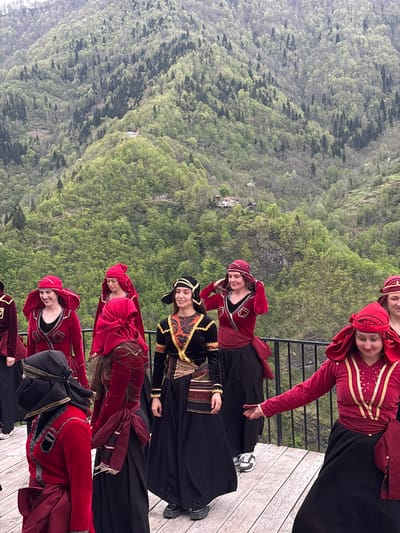 On a wooden platform overlooking a lush, mountainous landscape near Kveda Chkhutuneti, Georgia, a group of approximately six women stand in a staggered line, likely posing for a photo. All are wearing traditional Georgian attire consisting of long black dresses, and red and gold embellished velvet jackets. The women are smiling and appear relaxed. The backdrop shows a steep mountainside covered in dense green forest, dotted with small buildings. The weather appears overcast and hazy. A simple black metal railing separates the platform from the steep drop-off.