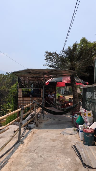 The image shows an outdoor area in Lâm Sơn, Vietnam. A small, makeshift structure with a roof and bamboo supports serves as a gathering place. Inside, several people sit at tables and chairs. A hammock is strung up nearby. The area is adjacent to a concrete walkway with scattered objects like a paint can and bags. A palm tree provides shade over the structure. Power lines are visible in the sky. A hill with trees is in the background.