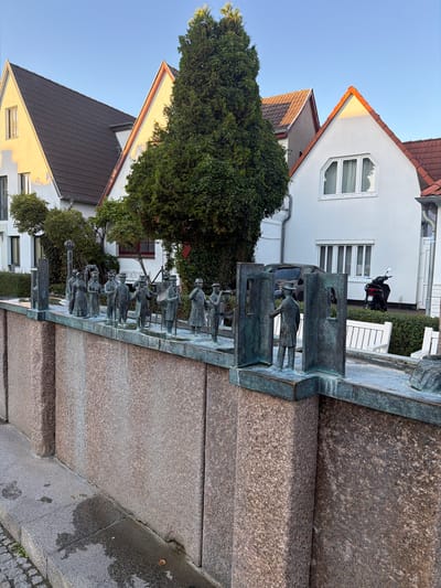 The image depicts an outdoor scene in Rostock, Germany, specifically at the "Zeitfigur" sculpture located on the Alter Strom. The sculpture, cast in bronze, features a line of figures, appearing to represent historical individuals, dressed in period clothing. The figures are arranged along a ledge that extends from a stone wall. The wall itself is constructed from large, rough-hewn granite blocks. In the background, modern buildings with white facades and red-tiled roofs are visible, along with a tall evergreen tree. The sky is clear and blue. A black scooter is parked near one of the buildings, and a dark-colored car is visible behind the sculpture. The ground in the foreground is paved with cobblestones and has wet patches, suggesting recent rain. The lighting indicates it is either early morning or late afternoon, with a warm glow on the building facades.