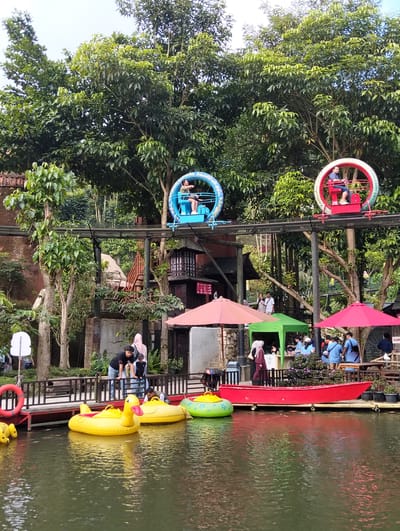 This media file captures a lively outdoor scene at what appears to be an amusement park or recreational area, likely in Gudangkahuripan, Indonesia, given the provided location context. The setting is characterized by lush greenery, with large trees dominating the background, suggesting a tropical or subtropical climate. The time of day appears to be daytime, with bright, diffused light, and the weather seems pleasant, possibly partly cloudy.

The foreground features a calm body of water, on which several inflatable boats are floating. Most prominent are two bright yellow duck-shaped inflatable boats, and to their right, a green and yellow inflatable boat. A long, striking red boat is docked alongside the water's edge. A red life preserver ring is visible on a wooden railing to the left.

In the mid-ground, there's a wooden deck and walkway, with a dark wooden railing separating it from the water. Several people are visible on and around this deck. Some are standing by the railing, while others are gathered under colorful umbrellas – a red one on the right, a pink one in the center, and a green canopy to its left. These areas seem to be service or rest spots.

Above the deck, an elevated track runs horizontally, supporting a unique amusement ride. Two open, circular carriages, one blue and one red, are shown on this track. Each carriage has a person seated inside, seemingly enjoying the ride. The blue carriage on the left has a person wearing a light-colored shirt, and the red carriage on the right has a person in a dark shirt and light-colored shorts.

Further in the background, there are rustic, wooden structures, including a two-story building with dark wood and white windows, reminiscent of traditional architecture. A wooden staircase leads up to it. A brick wall is visible to the far left.

The people in the scene appear to be engaged in various leisure activities, suggesting a recreational outing. The atmosphere seems relaxed and cheerful, typical of a park or tourist destination. There is some visible text on a sign near the red umbrella, reading "KASIR" which means "cashier" in Indonesian, indicating a point of sale. The overall impression is of a vibrant and enjoyable public space with a blend of natural beauty and man-made attractions.