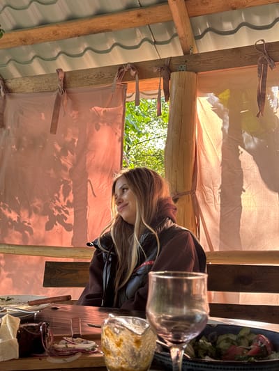 A photograph captures a woman with light brown hair sitting at a wooden table covered with food and a glass of wine, underneath a makeshift structure with a translucent, light peach-colored tarp roof. The tarp roof is secured to a wooden frame with tied ribbons. Visible through the frame are green leaves. The table is cluttered with dishes, including a salad. The woman is wearing a brown hooded jacket with a black leather overlay. There is no explicit mention or visual evidence linking the image directly to the city of Abzhanda, Abkhazia.