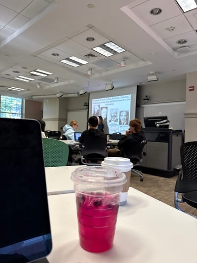 The image depicts a classroom scene within an academic institution in Wellesley, United States. In the immediate foreground, positioned on a white table, a clear plastic cup filled with a bright pink liquid containing dark, floating particles is visible, alongside a white disposable coffee cup with a brown protective sleeve. To the left, a portion of a dark-colored electronic device, likely a laptop, is partially in view.

The midground shows several individuals, primarily students, seated at white tables, facing a large projector screen. Most individuals are engaged with laptops. One student in a light blue hoodie is seated at a table with a laptop, another in a black hoodie is also at a laptop, and a third student with reddish-brown hair is observed looking towards the screen. A partially obscured individual, possibly an instructor, is visible near the projector screen on the left.

The background is dominated by a large white projection screen displaying a presentation slide. The slide's title reads "Context: Events, Ideas, Policies, Debates, Centering on the Role of Government in the Economy." Below the title, images of historical figures, including Adam Smith, Karl Marx, and John Maynard Keynes, are visible. To the right of the screen, a dark gray instructor's lectern or desk is present. The classroom features a dropped ceiling with square acoustic tiles, recessed fluorescent lighting, and HVAC vents. Large windows on the left side of the room offer a view of green foliage outside. The floor is carpeted.