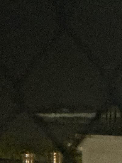 The image depicts a dark, grainy, and blurred scene at night in Ngũ Hành Sơn Ward, Vietnam. A prominent horizontal band of white and light grey blur extends across the mid-ground, consistent with breaking waves on a body of water, likely the ocean. Above this, the sky is uniformly dark. In the lower left foreground, several indistinct, warm-toned (orange/yellow) lights are visible, indicating artificial illumination from structures or streetlights. To the lower right, a portion of a light-colored, solid structure, possibly a building or wall, is partially visible. Faint, dark vertical blurs to the right of the wave line may represent distant structures or vegetation. No individuals, specific actions, or interactions are discernible.