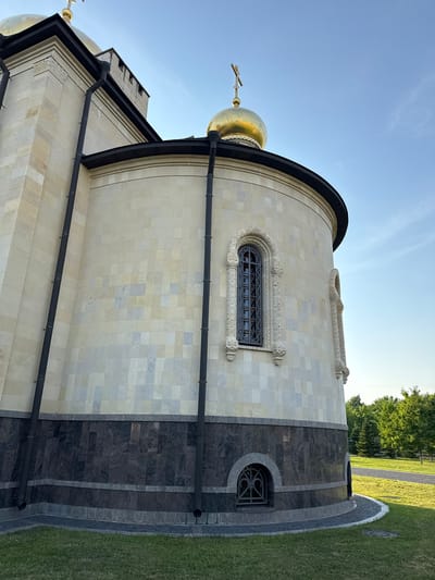 The image shows the exterior of a building, possibly a church, with a golden dome topped with a cross, visible against a clear blue sky. The building's facade is constructed of light-colored stone tiles arranged in a mosaic pattern. A dark-colored band runs around the lower portion of the building, above which is a section of dark granite with a lighter granite trim. Two black downspouts run vertically down the side of the building. There is a small arched window with a decorative metal grate on the lower part of the building. A paved path and manicured grass are visible in the foreground and to the right, with a line of trees in the background. This is the Church of the Assumption of the Blessed Virgin Mary in Kirishi, Russia.