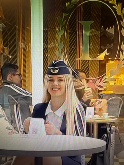 The image shows a woman with blonde hair wearing a dark blue hat with gold trim and a dark blue jacket over a white collared shirt. She is smiling and looking forward. She is sitting at a table in what appears to be an outdoor cafe. To her left, a person wearing glasses and a grey jacket with white stripes is seated. In the background, there is a wall with a pattern of diagonal lines and a large, ornate circular logo with a stylized "L" in the center. The scene is lit by natural light. The image quality is low, with visible pixelation and blurriness. No specific details about the location in Chorzów, Poland are discernible from the image.